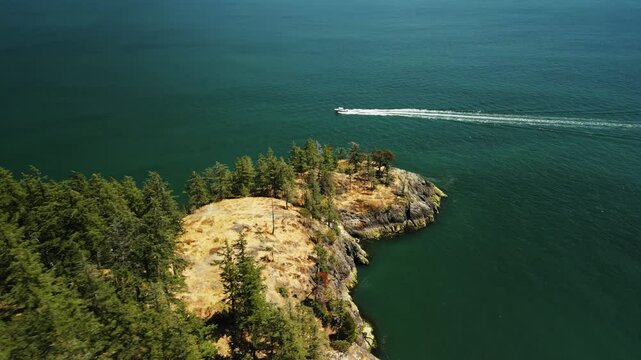drone shot as speedboat sails around the small peninsula of a small tropical island covered in sandy rocks and trees surrounded by clear blue water on a sunny day