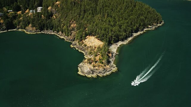 Aerial shot of speed boat sailing around a rocky peninsula on Bowen Island in Vancouver with hilly forests and surrounded by tropical blue ocean water on a sunny summer day