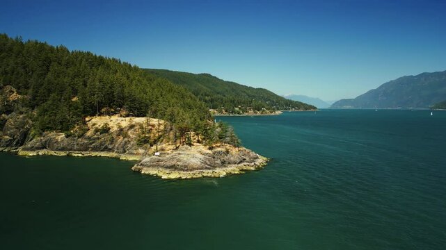 Drone orbit around small peninsula on local Bowen Island surrounded by tropical clear blue water revealing the long winding coastline in remote part of Canada with mountains in the background