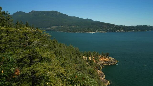 Drone flies around tree line of an island forest to reveal the waters edge with rocky peninsula surrounded by tropical blue water and a mountain range in the background on sunny day