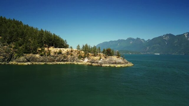 Drone shot flying around a rocky peninsula located on Bowen Island surrounded by trees, snow capped mountains, cliff edges and clear blue tropical water for swimming on a sunny day in Vancouver