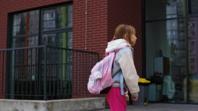 A young girl, around 7-9 years old, is joyfully skipping toward school, seen from the side, wearing a grey and white varsity jacket, pink pants, and carrying a large pink backpack with blue straps.