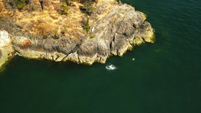 Drone shot capturing friends jumping off a small cliff side at the edge of an island into the tropical blue green water on a sunny summer day from yellow sandy stone rocks