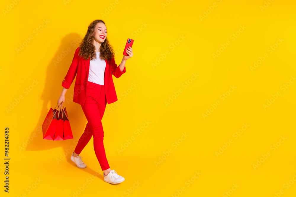 Wall mural Full size photo of pretty young woman use phone walk empty space wear red suit isolated on yellow color background