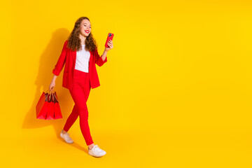 Full size photo of pretty young woman use phone walk empty space wear red suit isolated on yellow color background