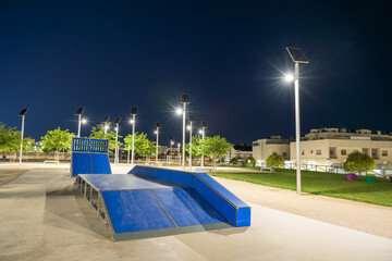 Skate park with ramps and rails in a clean urban environment, illuminated at night by several solar-powered street lights, highlighting the sustainable energy infrastructure in public spaces. Spain