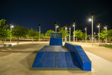 Modern skate park with solar-powered lighting in a residential area near a housing estate at night. Solar-powered street lights provide energy-efficient lighting during blue hour