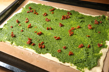 Puff pastry dough topped with homemade green pesto and dried tomatoes on baking paper and a baking...