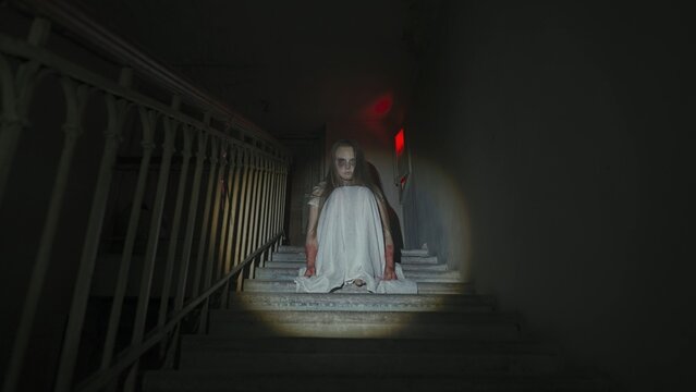 Creepy ghost woman in white dress and scary makeup alone on staircase in dark haunted house lighted by flashlight. Halloween horror advertisement concept.