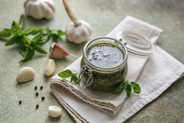 Top view of homemade pesto sauce in glass jar, basil, garlic and pine nuts on table. Preparing...