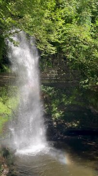 Glencar Waterfall in Leitrim, Ireland with a rainbow mist, surrounded by lush greenery and rocky pool.