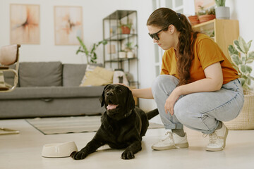 Young woman petting excited black Labrador while he lying on floor