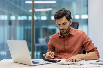 Obraz premium Young male entrepreneur working in office using calculator and laptop. Serious and focused on financial calculations, seated indoors at modern workplace. Represents business and productivity