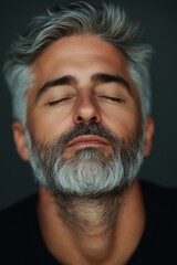 A contemplative man with gray hair and beard relaxes with his eyes closed against a dark background in an intimate setting