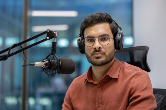 Serious male broadcaster with glasses and headphones sits in studio recording podcast. Professional setting conveys focus and expertise. Capturing essence of modern media and communication.