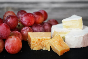 A bunch of large pink grapes next to Brie and Parmesan cheese on a black tray on a gray wooden background