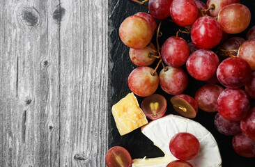 A bunch of large pink grapes next to Brie and Parmesan cheese on a black tray on a gray wooden background