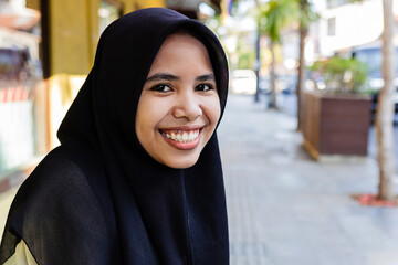 Portrait of happy young Asian Muslim woman wearing hijab smiling at camera sitting at city street. Head shot of beautiful indonesian student girl outside.