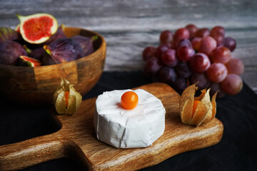 Ripe figs next to Brie cheese on a wooden board, pink grapes and Physalis fruits on a dark background