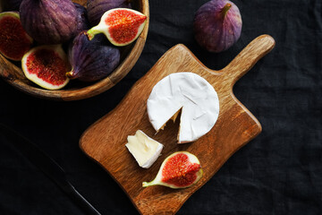 Ripe figs in a wooden bowl next to Brie cheese on a wooden board on a dark background