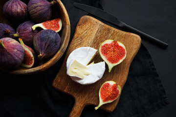 Ripe figs in a wooden bowl next to Brie cheese on a wooden board on a dark background