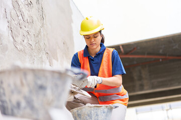Skilled young female bricklayer with trowel taking cement from bucket while plastering wall at...