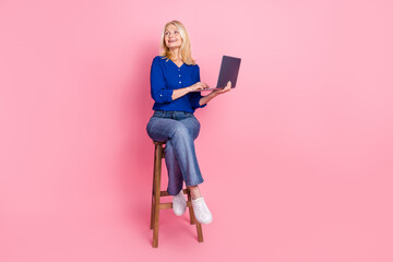 Full length photo of lovely pensioner lady sit chair hold netbook look empty space dressed blue garment isolated on pink color background