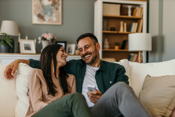 Obraz premium Loving modern couple sitting on the sofa, man holding a remote controller and watching a TV together