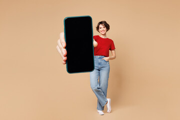 Full body young happy woman she wear red t-shirt casual clothes hold in hand use mobile cell phone with blank screen workspace area isolated on plain pastel light beige background. Lifestyle concept.