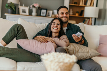 Diverse couple, watching tv and remote on living room couch with smile, hug or relax with click for...