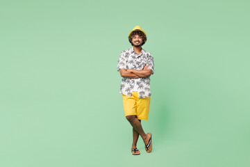 Full body young smiling happy Indian man wear beach shirt hat rest near hotel pool hold hands crossed folded look camera isolated on plain green color background. Summer vacation sea sun tan concept.