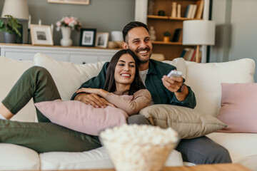 Diverse couple, watching tv and remote on living room couch with smile, hug or relax with click for choice, show or film