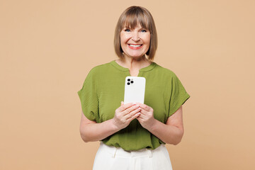 Elderly smiling happy blonde woman 50s years old she wearing green shirt casual clothes holding in hand use mobile cell phone isolated on plain pastel light beige background studio. Lifestyle concept.