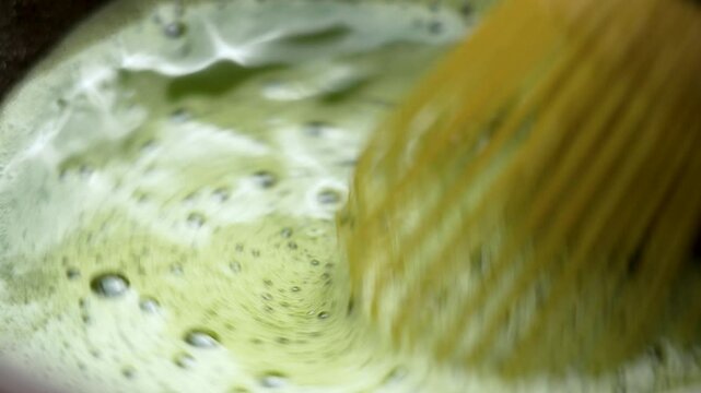 Whisking Japanese green matcha tea in a bowl with a bamboo whisk. Preparing traditional hot tonic drink. Macro shot