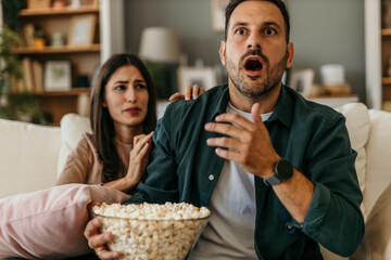 An upset adult couple watching a scary movie on television