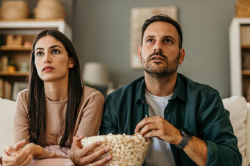 Young married couple, man and woman, sitting at home on sofa, eating popcorn and watching scary horror movie or dramatic sports games action or drama film. Simple living concept, weekend activities.