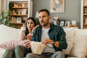Multiracial husband and wife eating popcorn from a bowl and watching drama movie while chilling on...