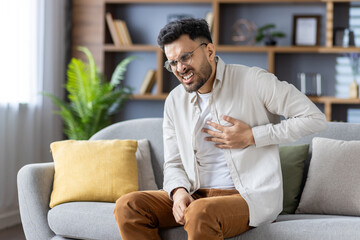 Young man experiencing chest pain sitting on couch expressing discomfort with hand on chest