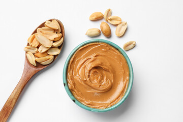 Tasty peanut butter in bowl and spoon with groundnuts on white table, flat lay
