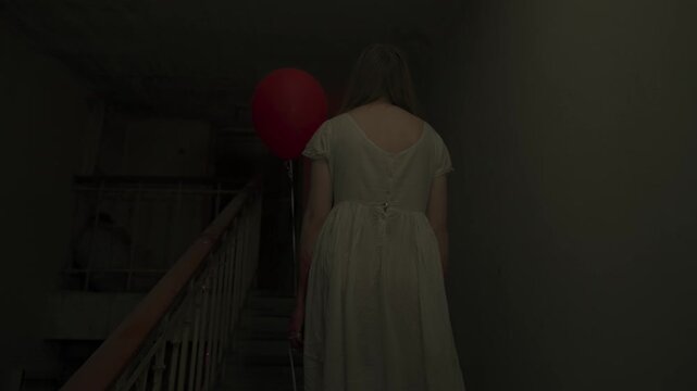 Creepy ghost woman in white dress and scary makeup walking up alone on staircase in dark haunted house holding red balloon. Halloween horror advertisement concept