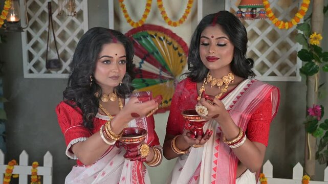 Two Indian women, draped in traditional sarees and adorned with gold jewellery, holding dhunachi in hands in front of a beautifully decorated house