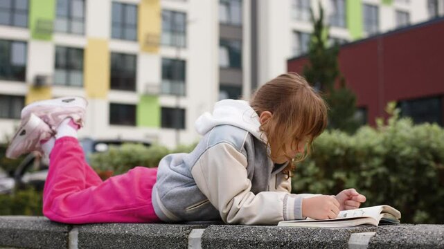 A young girl, around 7-9 years old, is lying on her stomach outdoors, completely focused on reading a book. She is wearing a grey and white varsity jacket, bright pink pants, and pink sneakers.