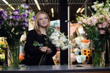 Fototapeta premium Happy young woman florist in suit collecting bouquet at workplace in flower shop