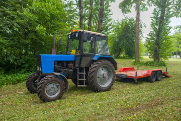 A blue tractor is parked in a field next to a red trailer. The tractor is surrounded by trees and grass