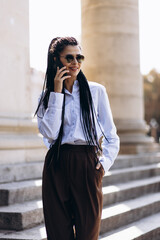 Fototapeta premium Young woman with braided hair walking downstairs of a beautiful architecture building and talking on the phone