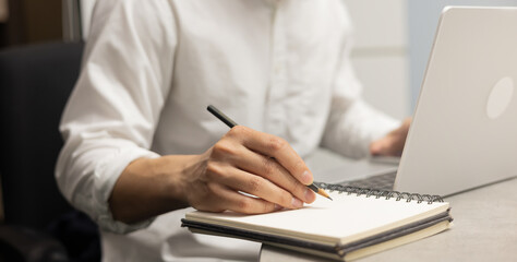 close up businessman hand writing content or somethings on notebook with using laptop at wooden table at home office for people and lifestyle concept