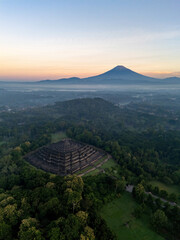 Borobudur - world's largest Buddhist Temple in Magelang, Indonesia dark before dawn sunrise (sun rising on horizon) with valley, trees, town in foreground, Mount Sumbing Gugung Sumbing in background.