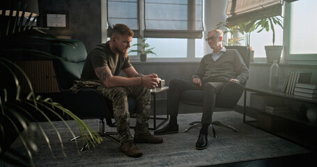 Depressed military man with PTSD sits in chair, talks to specialist on counseling session. Senior psychotherapist listens and communicates with patient during therapy. Post traumatic stress disorder.