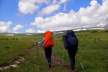 Two girls with backpacks are walking along a trail in the mountains