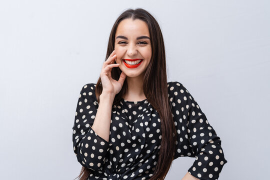 Cheerful woman with long hair smiling in a dotted blouse. A woman with long hair and bright red lipstick smiles widely while wearing a black and white dotted blouse against a light background.